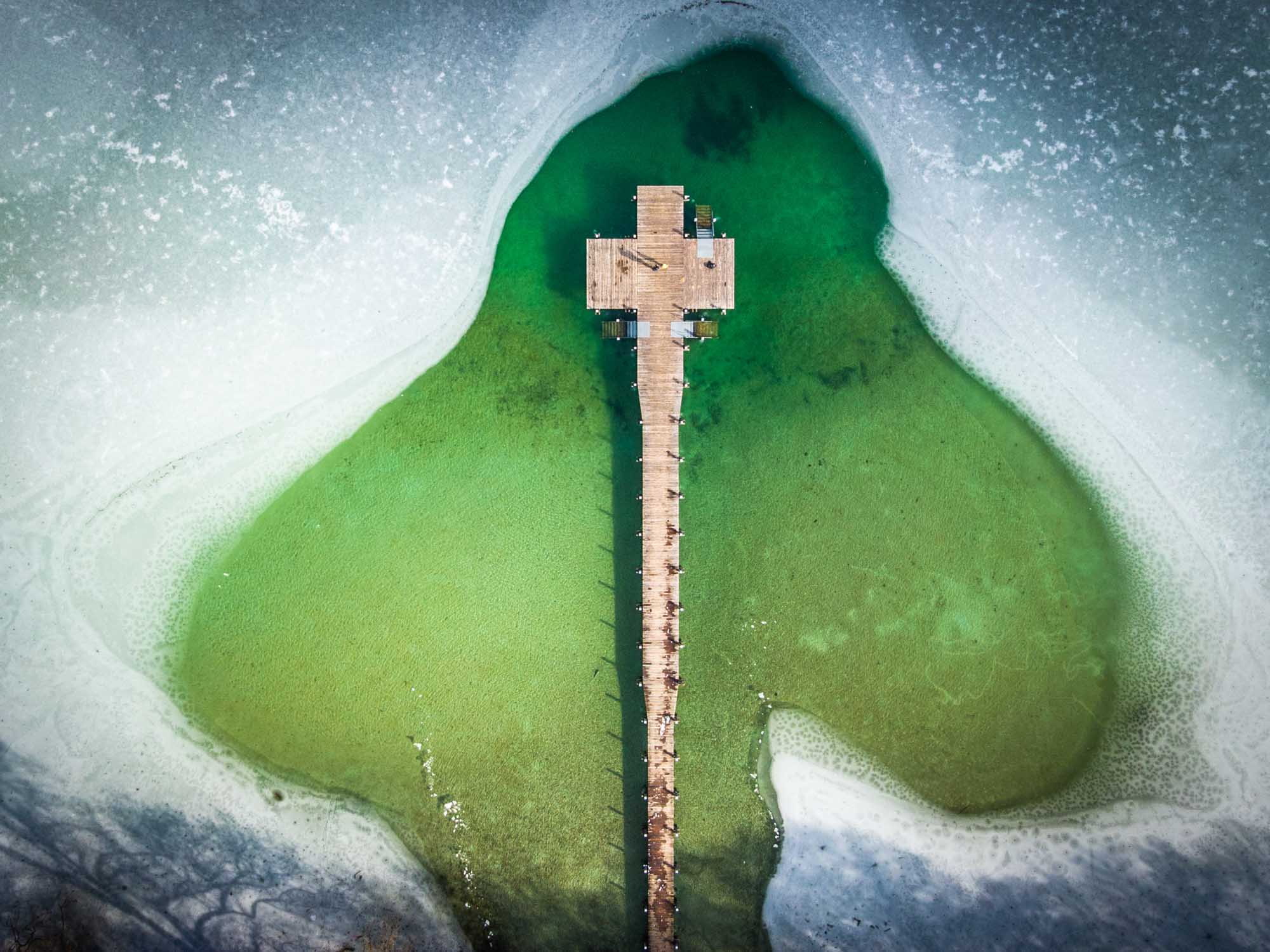 Aerial view of a wooden pier over a frozen lake with clear green water and ice surrounding it.