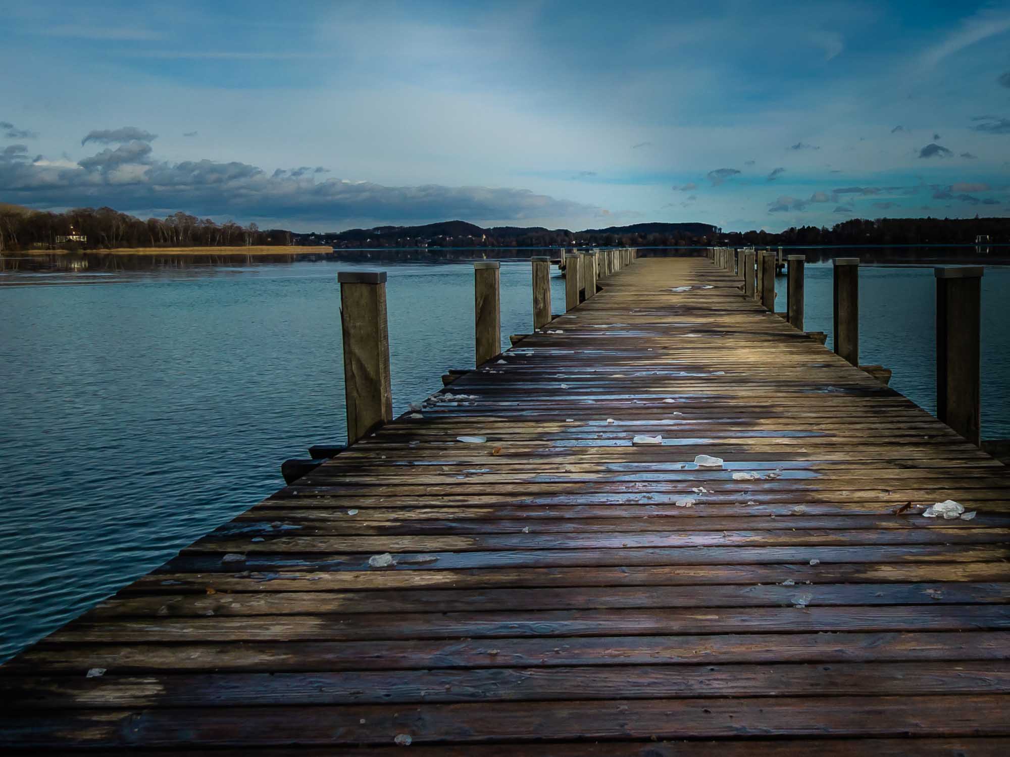 Wooden pier stretching into a calm lake under a clear blue sky, surrounded by tranquil water and distant trees.