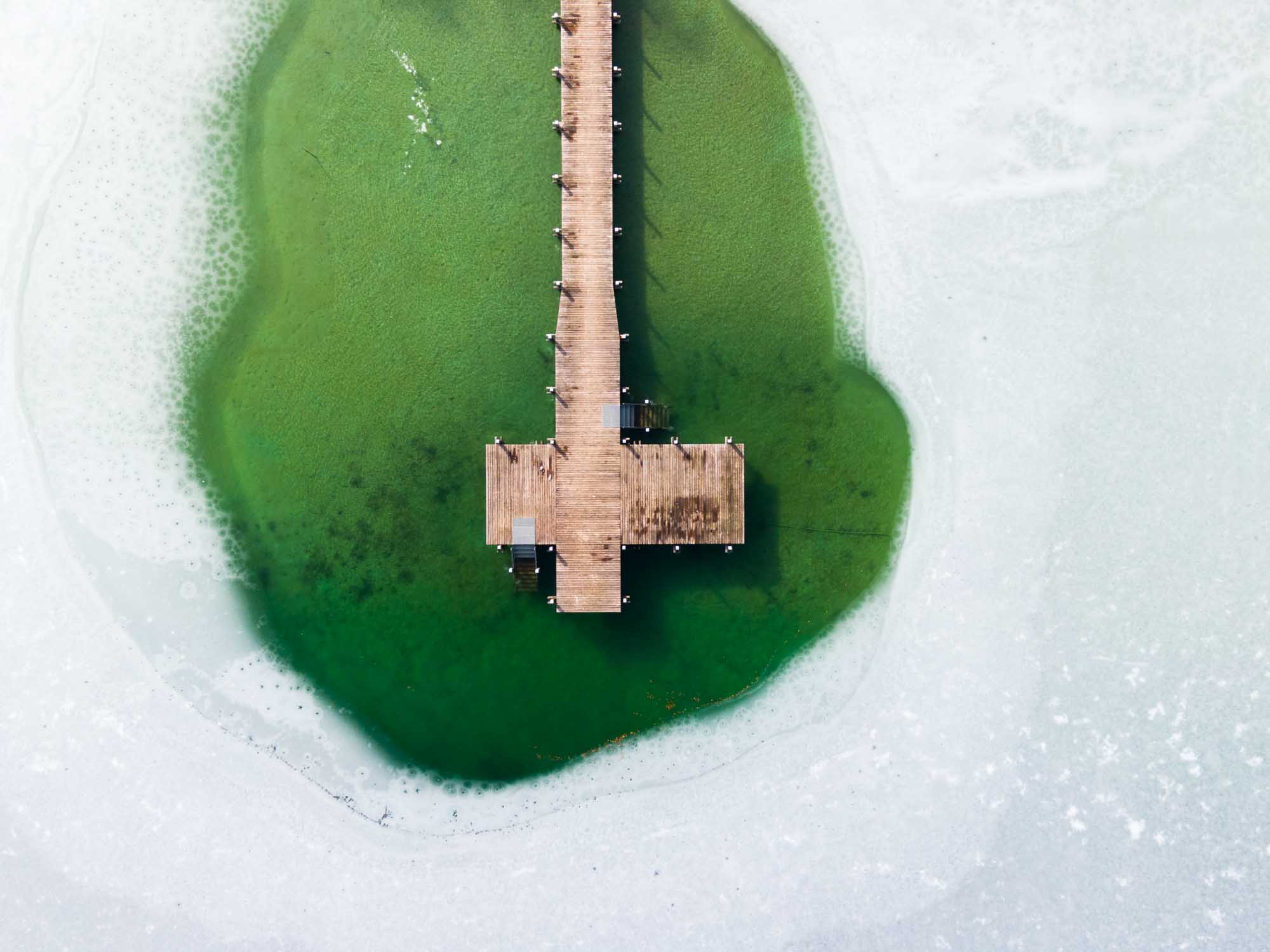 Aerial view of a wooden pier extending over green water surrounded by ice.