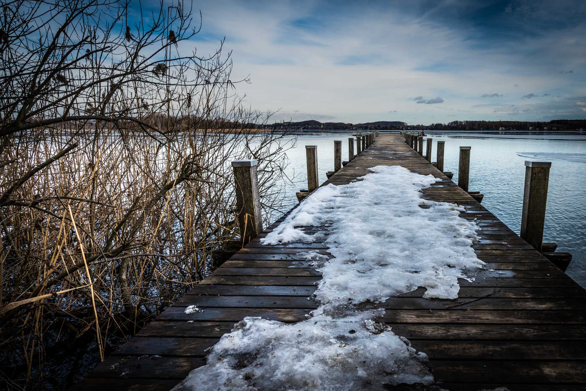 Snow-covered wooden pier extending into a calm lake, surrounded by bare winter trees under a cloudy sky.