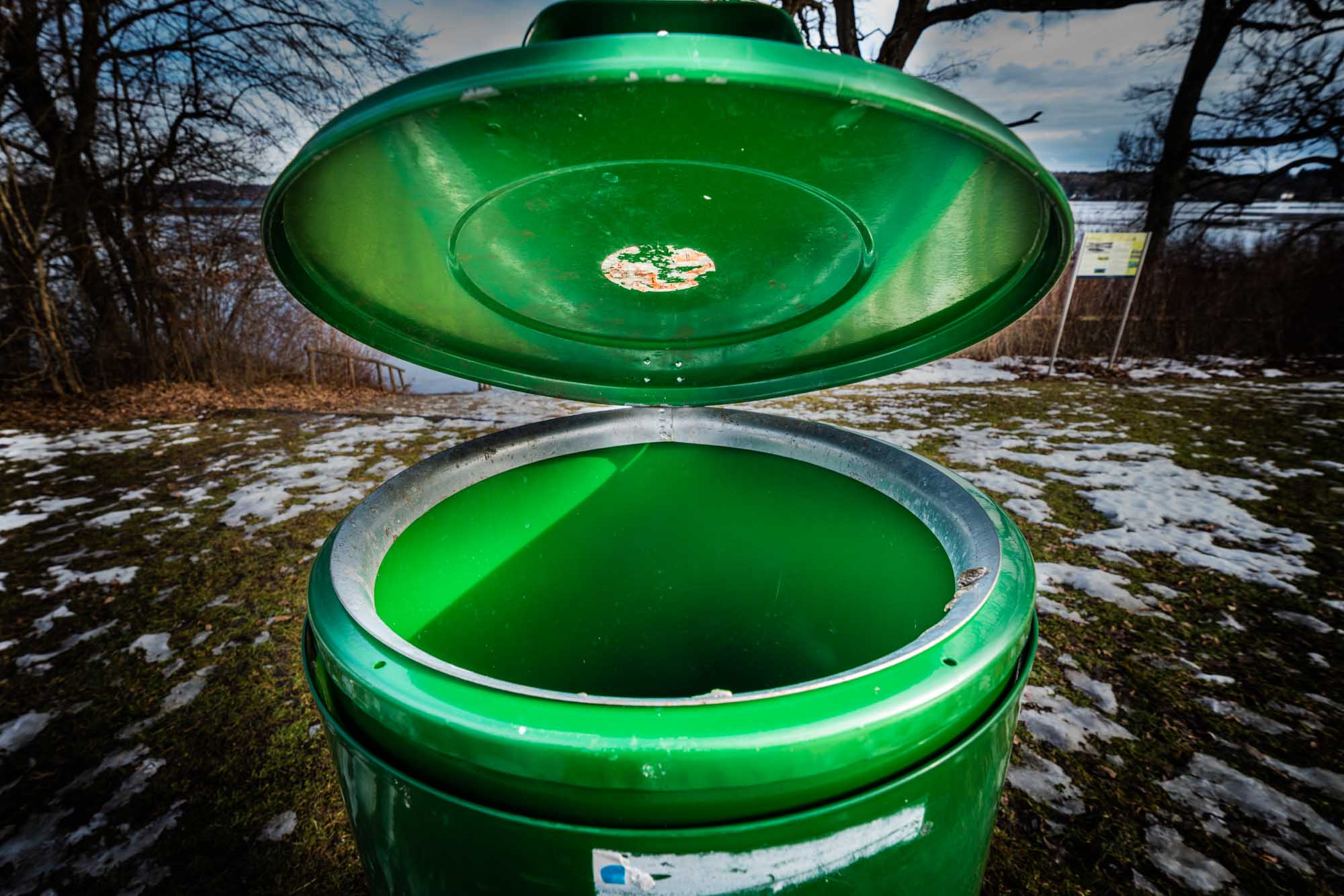 Open green trash can in a snowy park with barren trees and lake in background under cloudy sky.