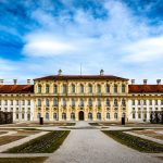 Elegant baroque palace under partly cloudy sky, surrounded by a formal garden and pathways.