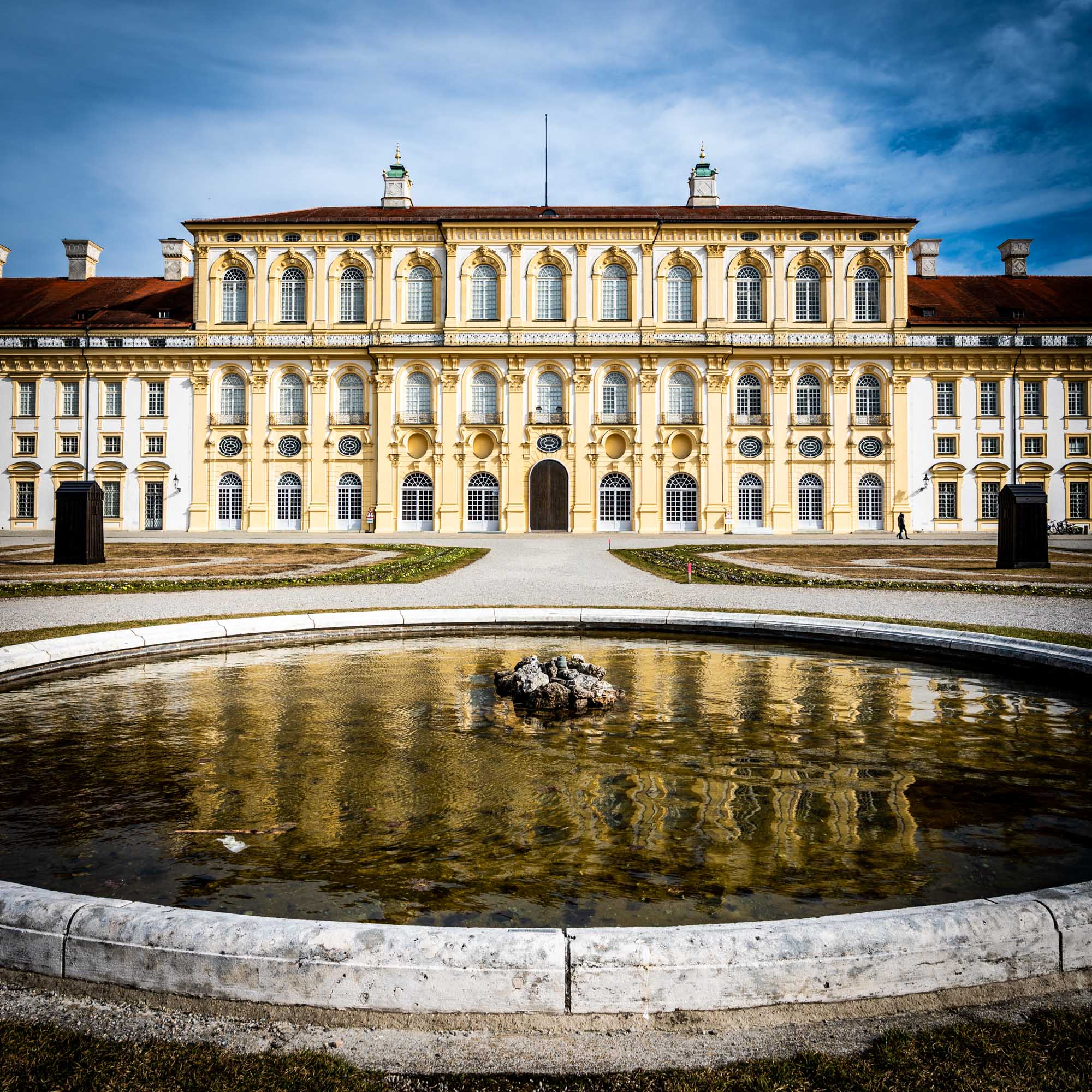 Baroque palace with ornate windows and a central entrance, reflected in a foreground fountain under a blue sky.