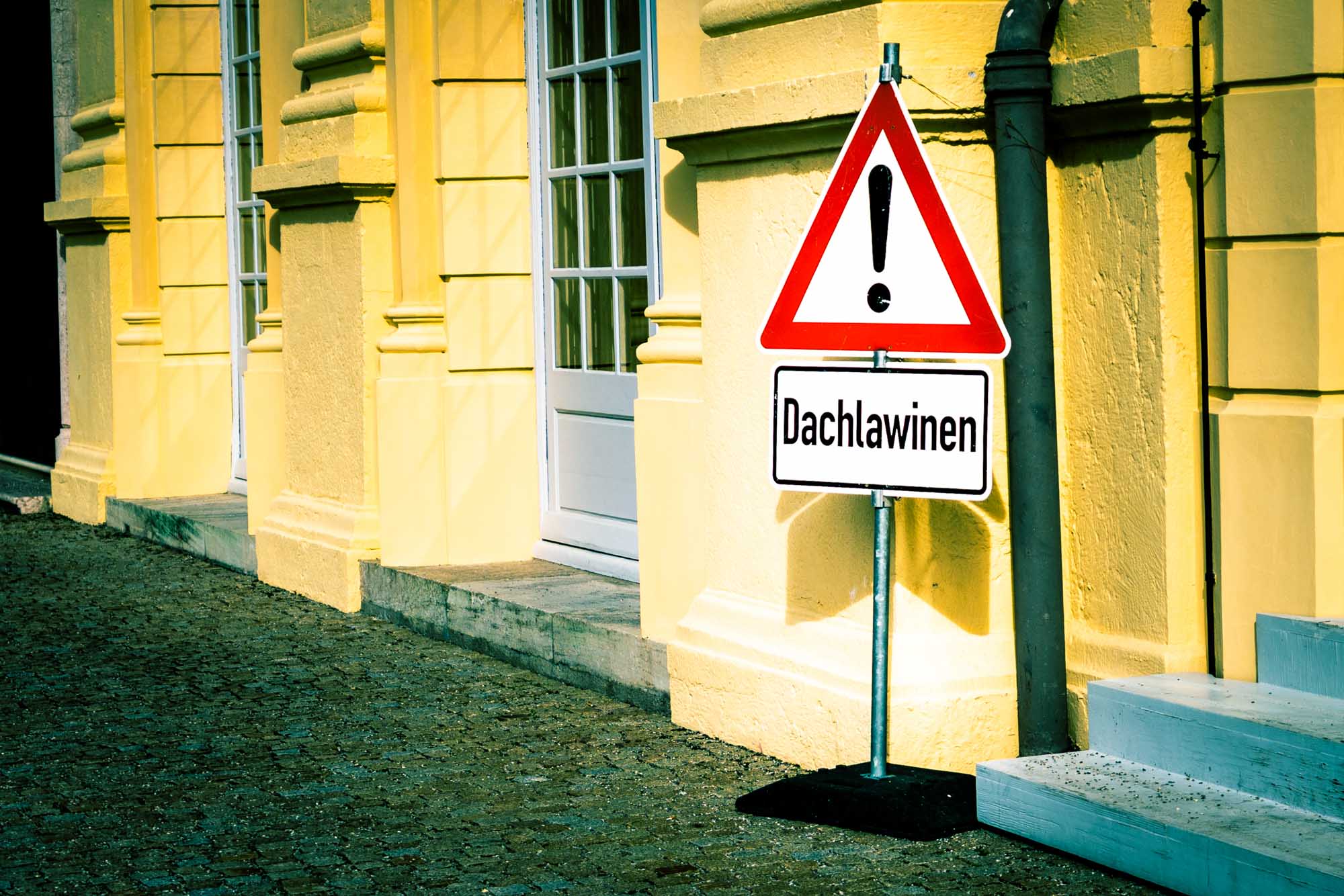 Warning sign for roof avalanches (Dachlawinen) against yellow building with glass doors.