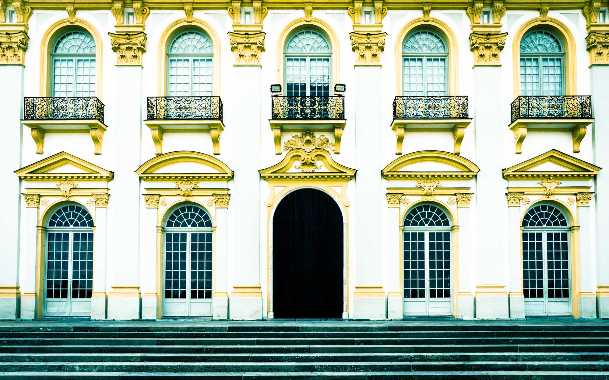 Elegant baroque architecture with arched windows, ornate balconies, and yellow accents above stone steps.