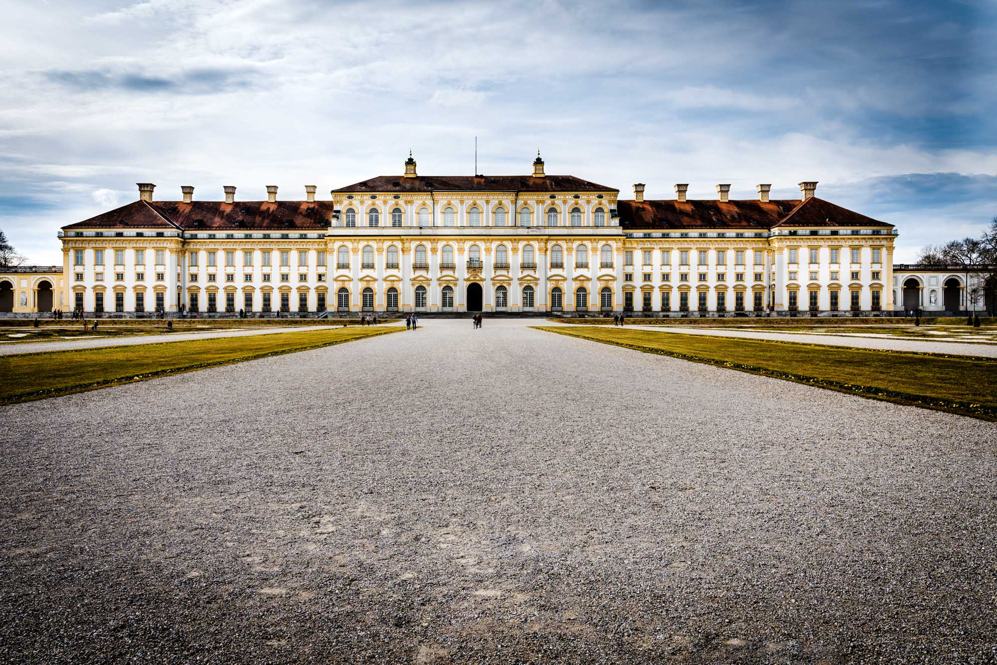 Grand historic palace with symmetrical architecture, central entrance, and expansive gravel pathway under a cloudy sky.