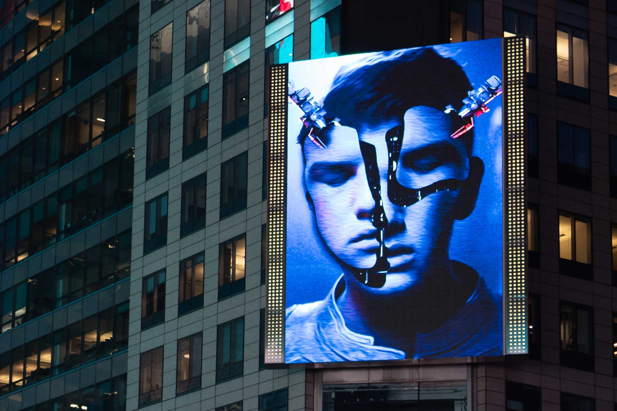Digital billboard on building displays a split-face artwork with blue tones and robotic elements at night.