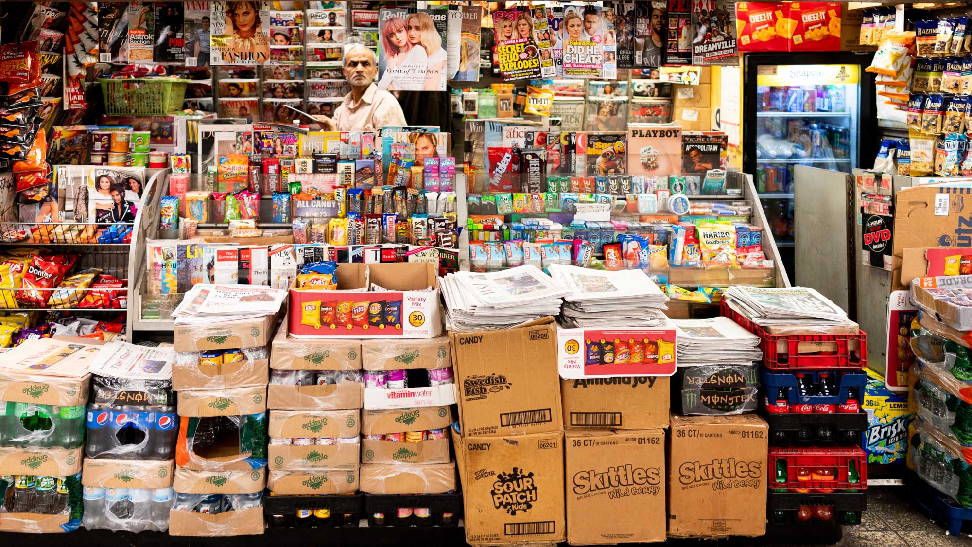 Bustling newsstand with magazines, snacks, and drinks. Stacked boxes and newspapers fill the busy space.