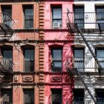 Colorful urban building facade with red, pink, white, and blue sections, featuring black fire escapes and window AC units.