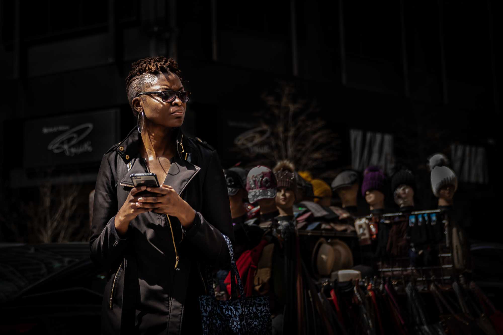 Person in sunglasses holding phone, standing by outdoor street market with hats and mannequins.