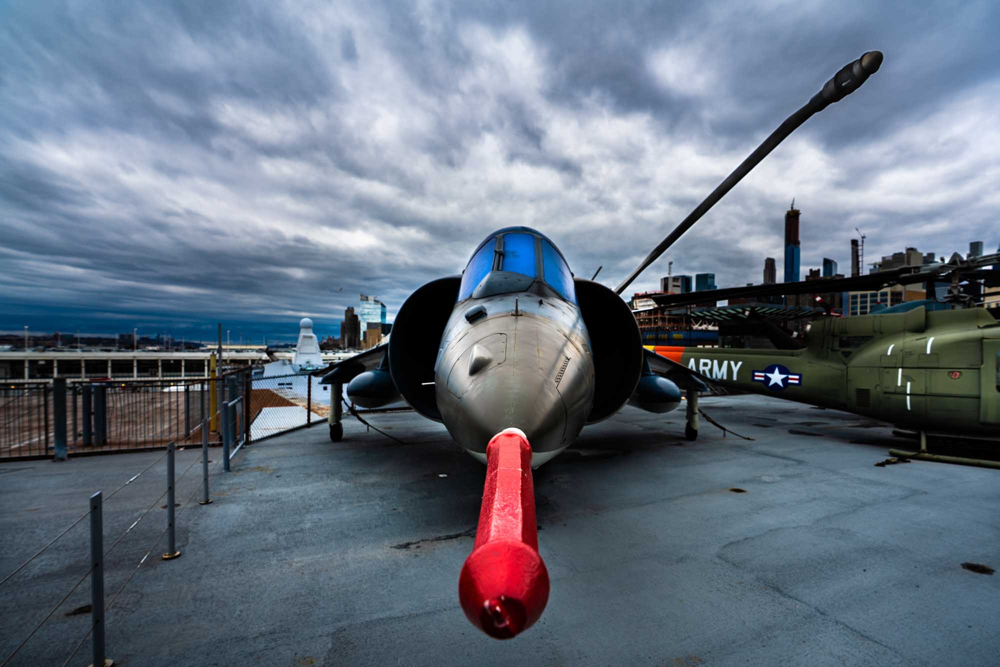 Military jet with red nose cone on aircraft carrier deck, cloudy sky, city skyline in background.