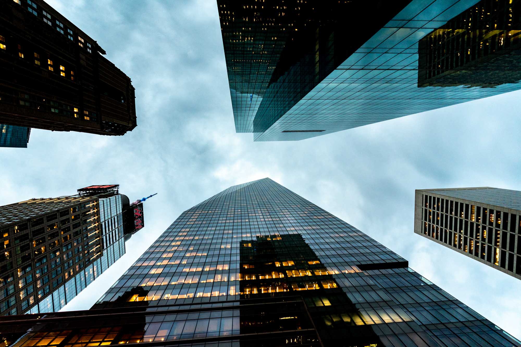 Looking up at tall skyscrapers with glass facades under a cloudy sky, creating a modern cityscape view.