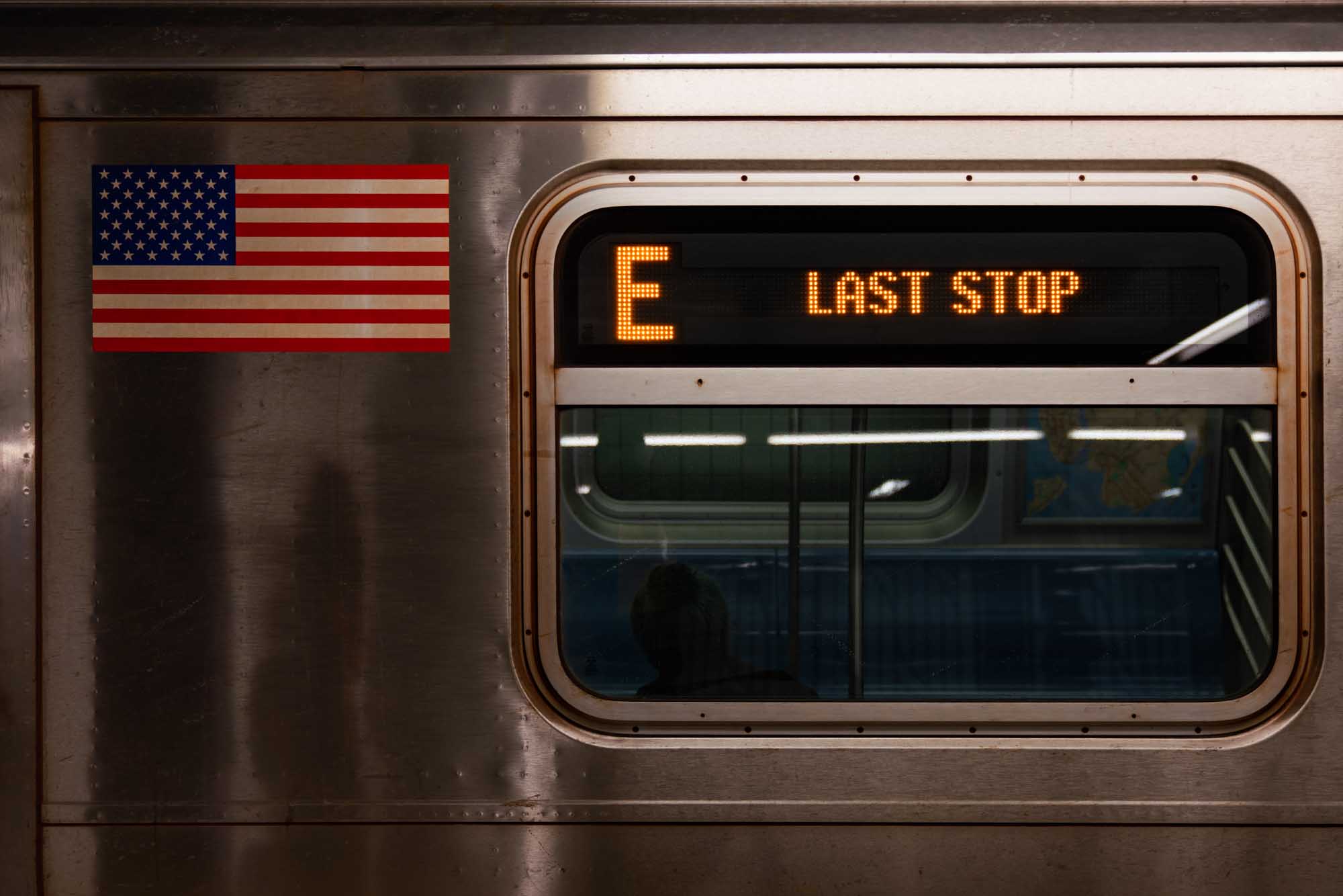 Subway train with E Last Stop sign and American flag on metallic exterior in dim light.