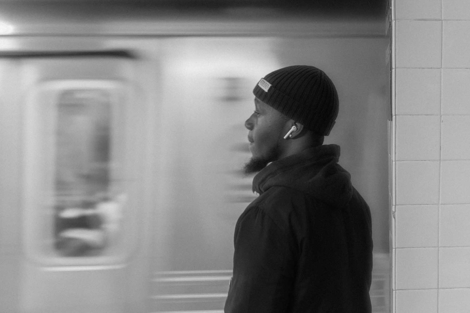 Man in beanie and earbuds waits on subway platform as train passes by, black-and-white image.