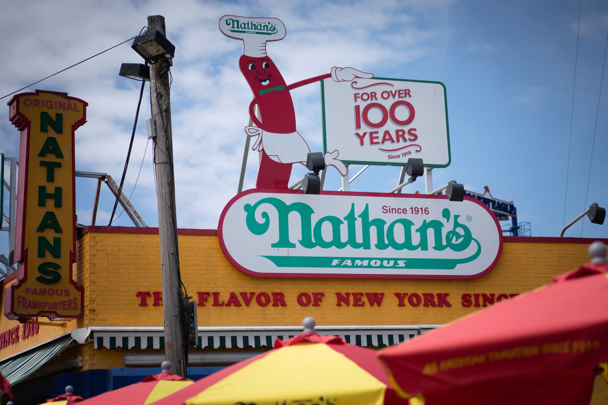 Nathan's Famous hot dog restaurant with vintage signs celebrating over 100 years at Coney Island, surrounded by umbrellas.
