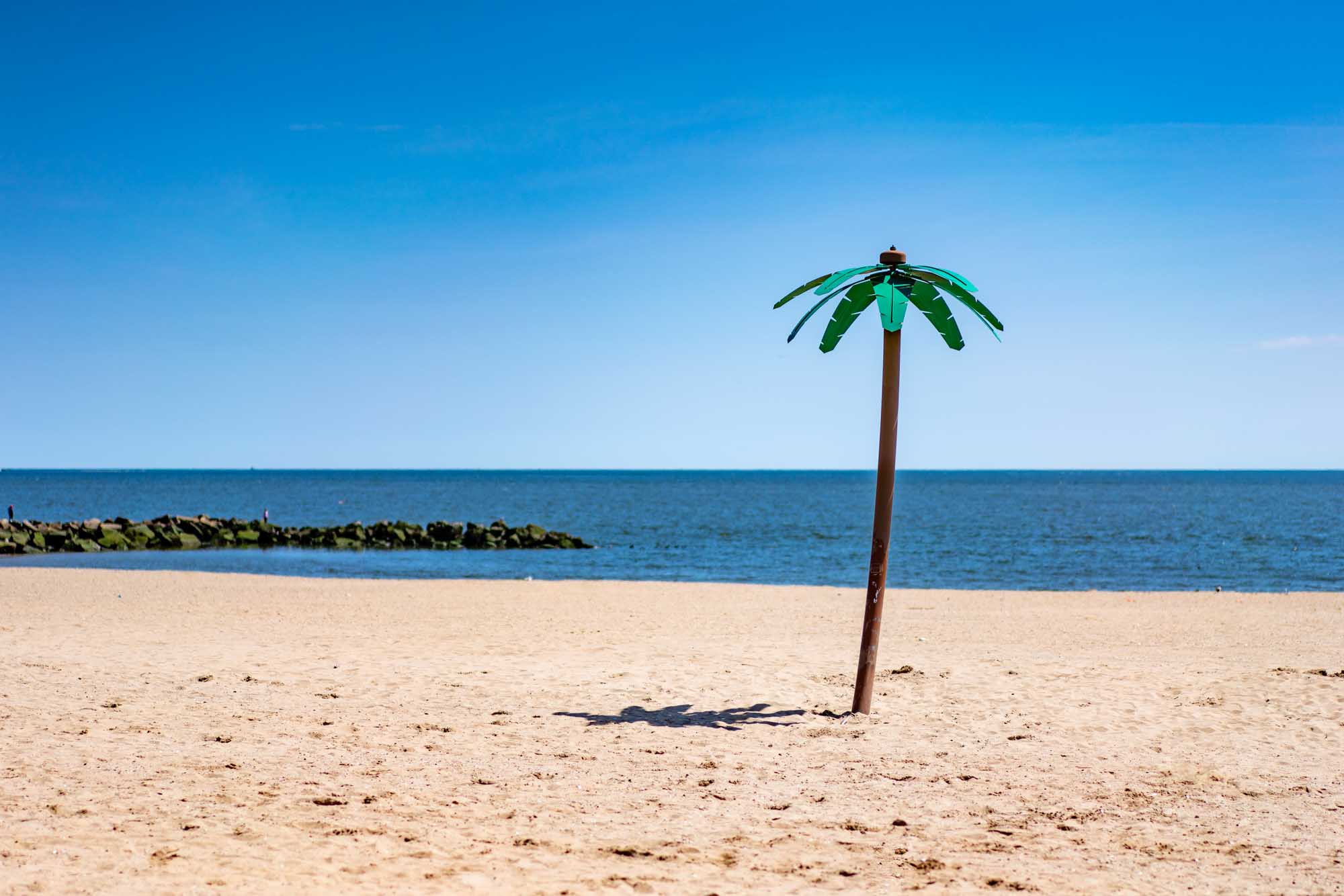 Artificial palm tree on a sandy beach with calm sea and a clear blue sky background.