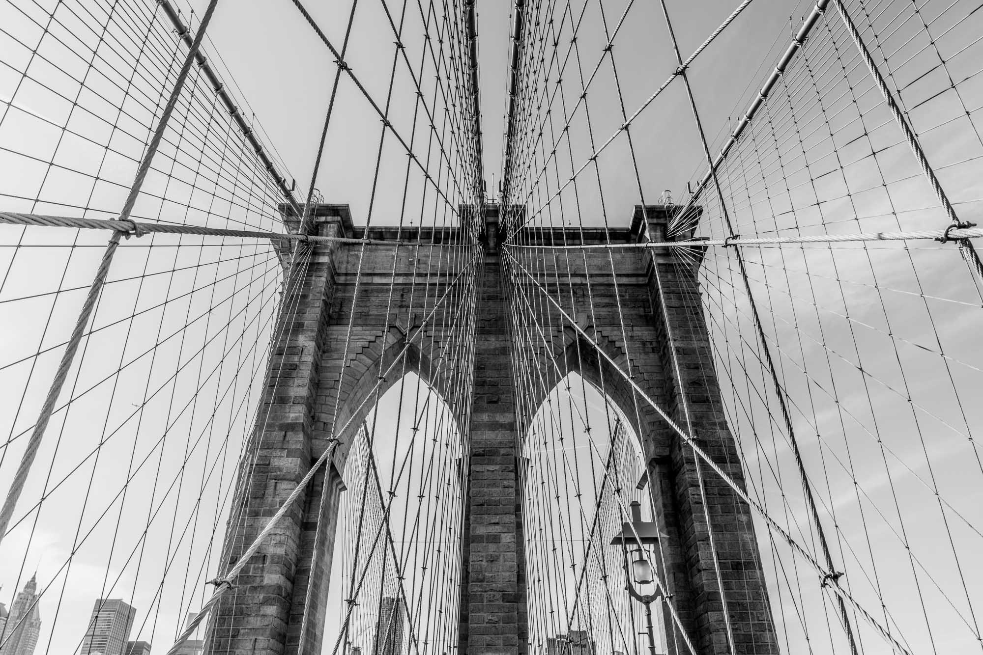 Black and white view of Brooklyn Bridge cables and arches against the sky, New York City skyline faintly visible.