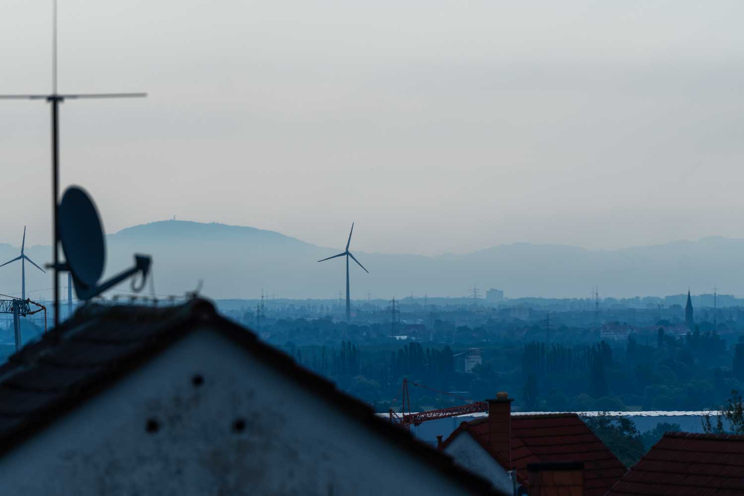 Rooftop view with wind turbines and distant hills under a cloudy sky, with blurred antennas in the foreground.