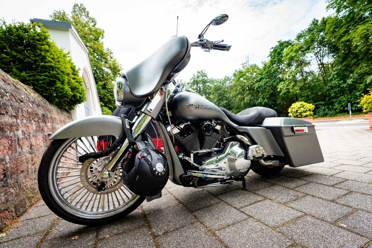 Low-angle view of a gray Harley-Davidson motorcycle parked on a paved area with trees and a white wall in the background.