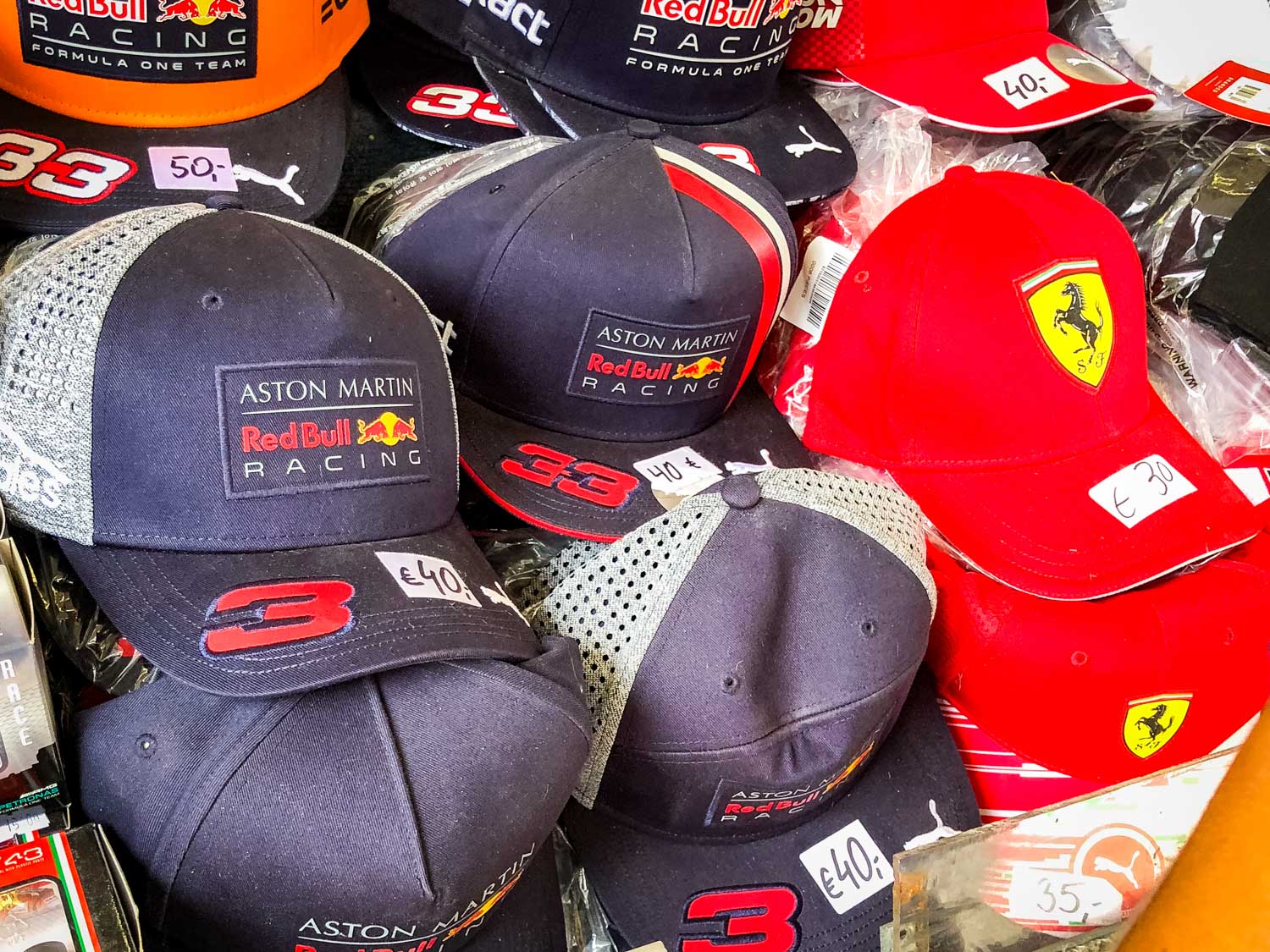 Assorted Red Bull and Ferrari branded caps for sale, priced with tags, showcasing motorsport merchandise.