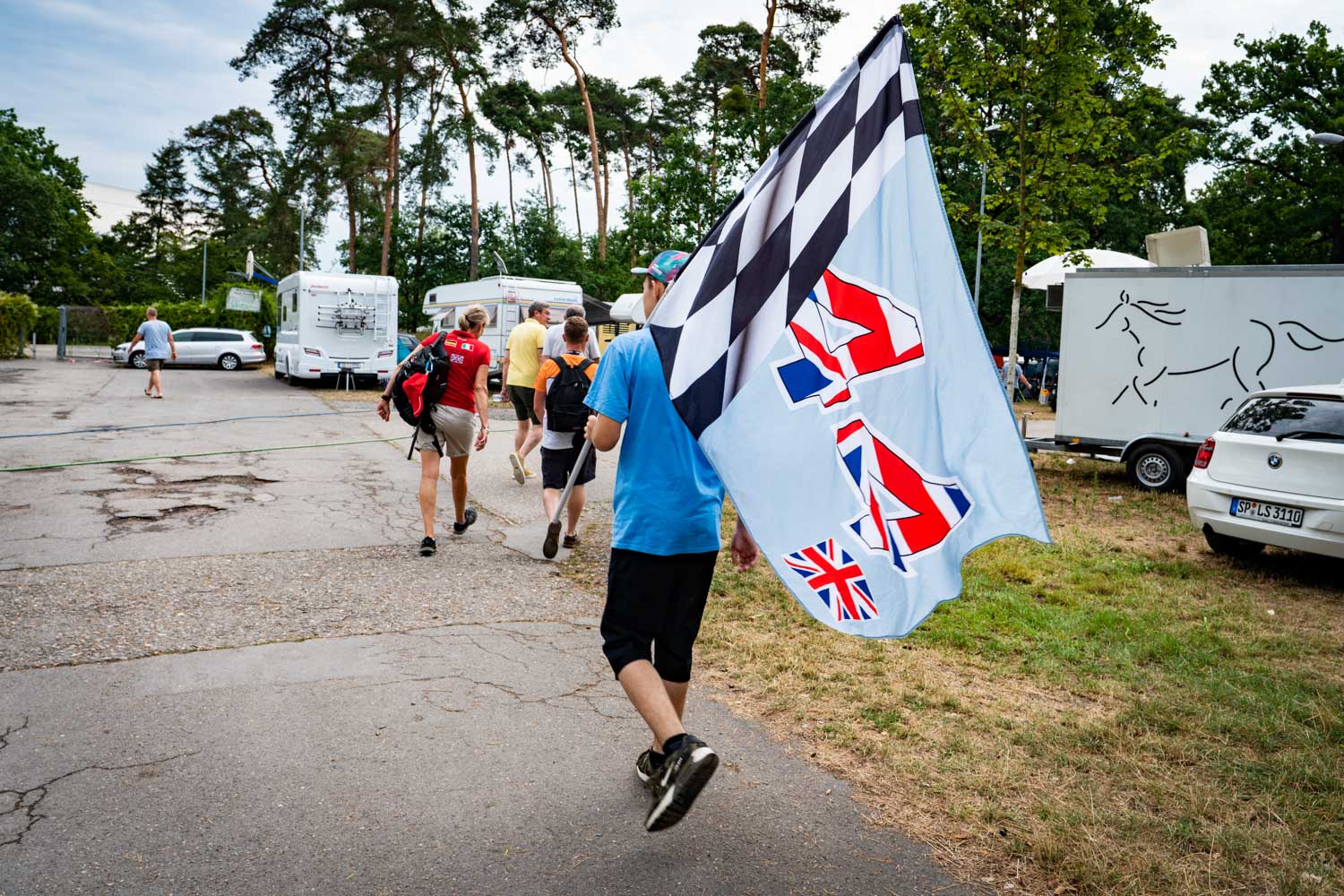 Person carrying a British flag with checkered design in park, surrounded by trees, campers, and parked cars.