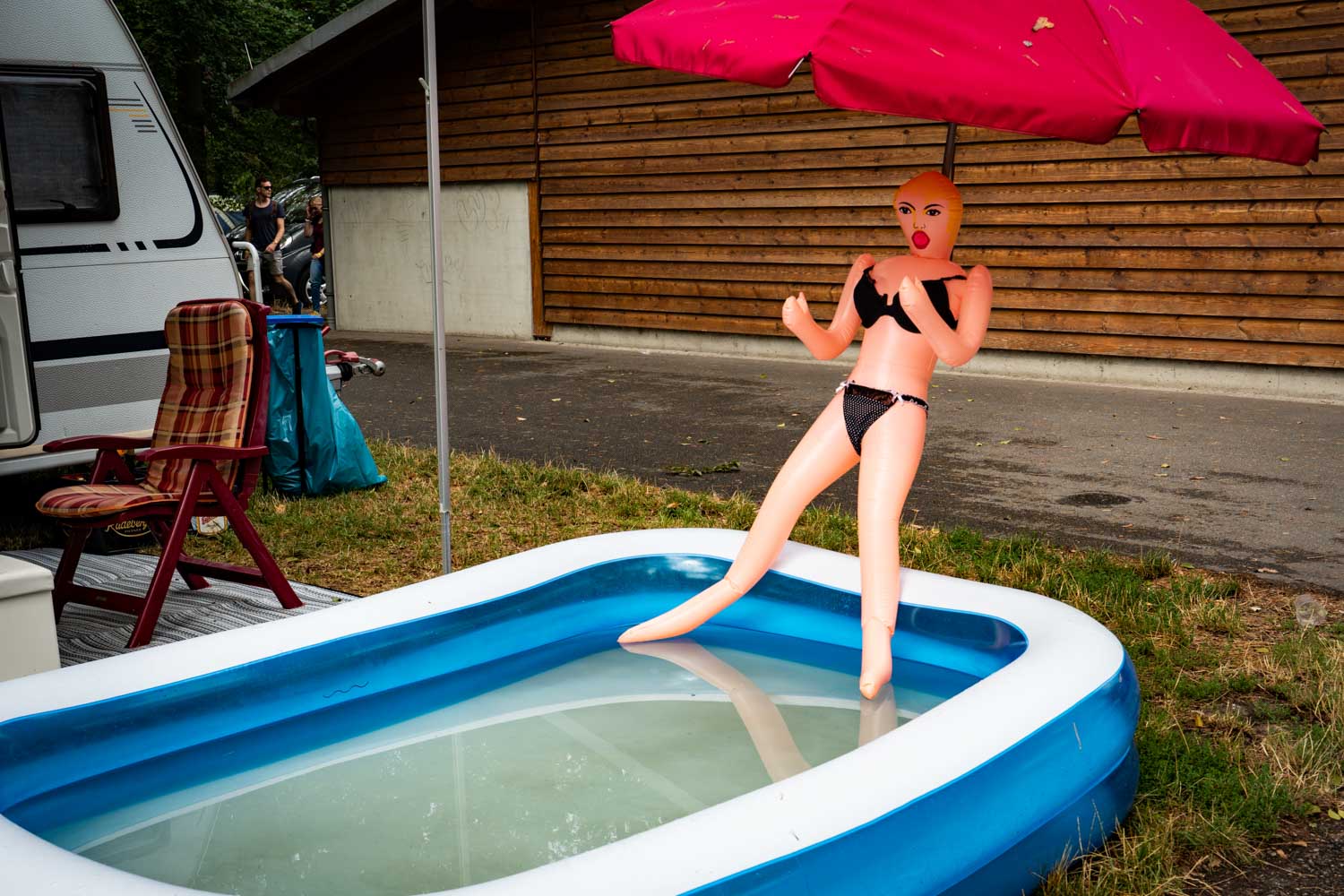 Inflatable doll in bikini by pool under red umbrella at campsite with trailer and wooden cabin in background.