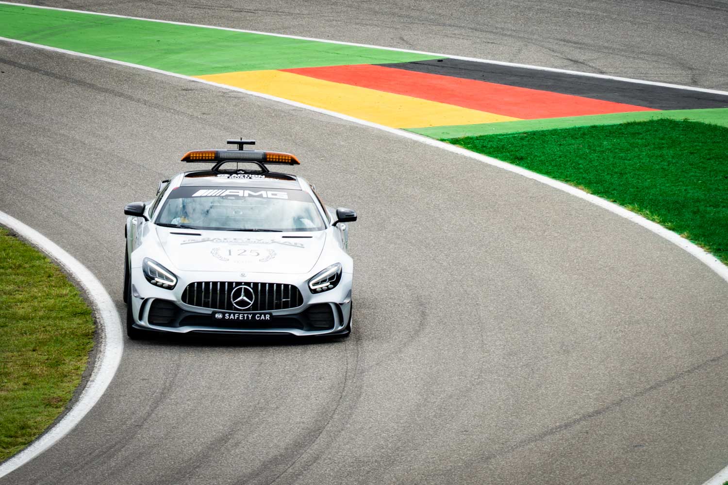 Silver Mercedes AMG safety car on a racetrack turn with green grass and colored kerbs.