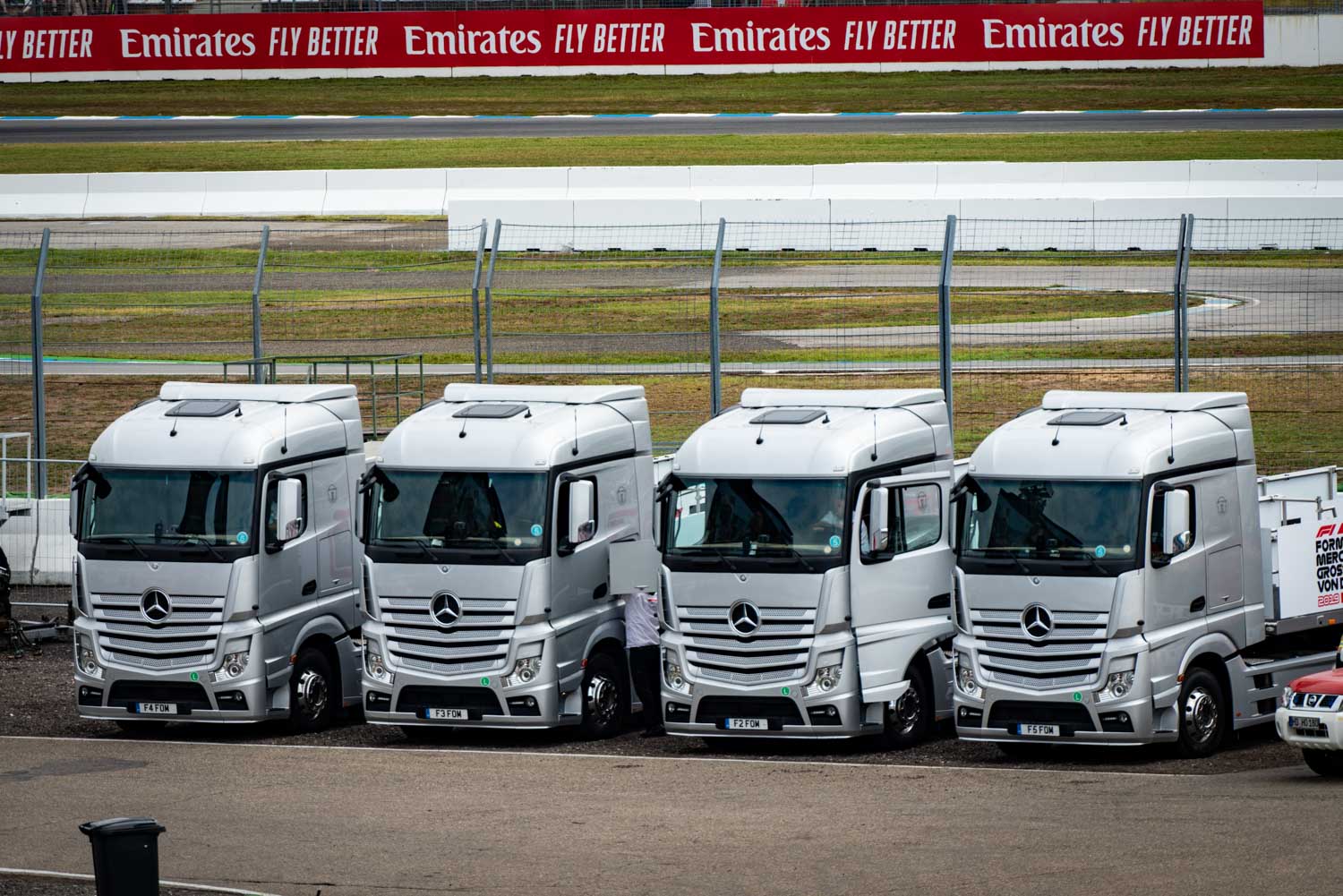 Four silver Mercedes-Benz trucks parked at a racetrack with Emirates advertising in the background.