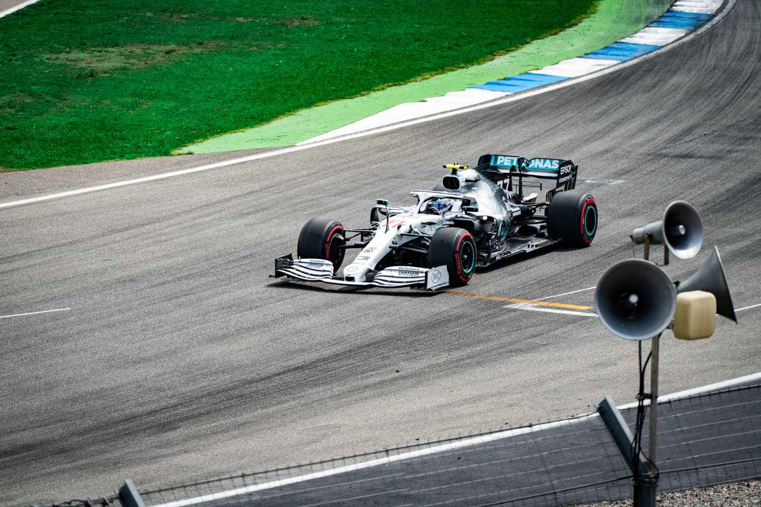 Silver Formula 1 car racing on track with green grass and megaphones in the background.