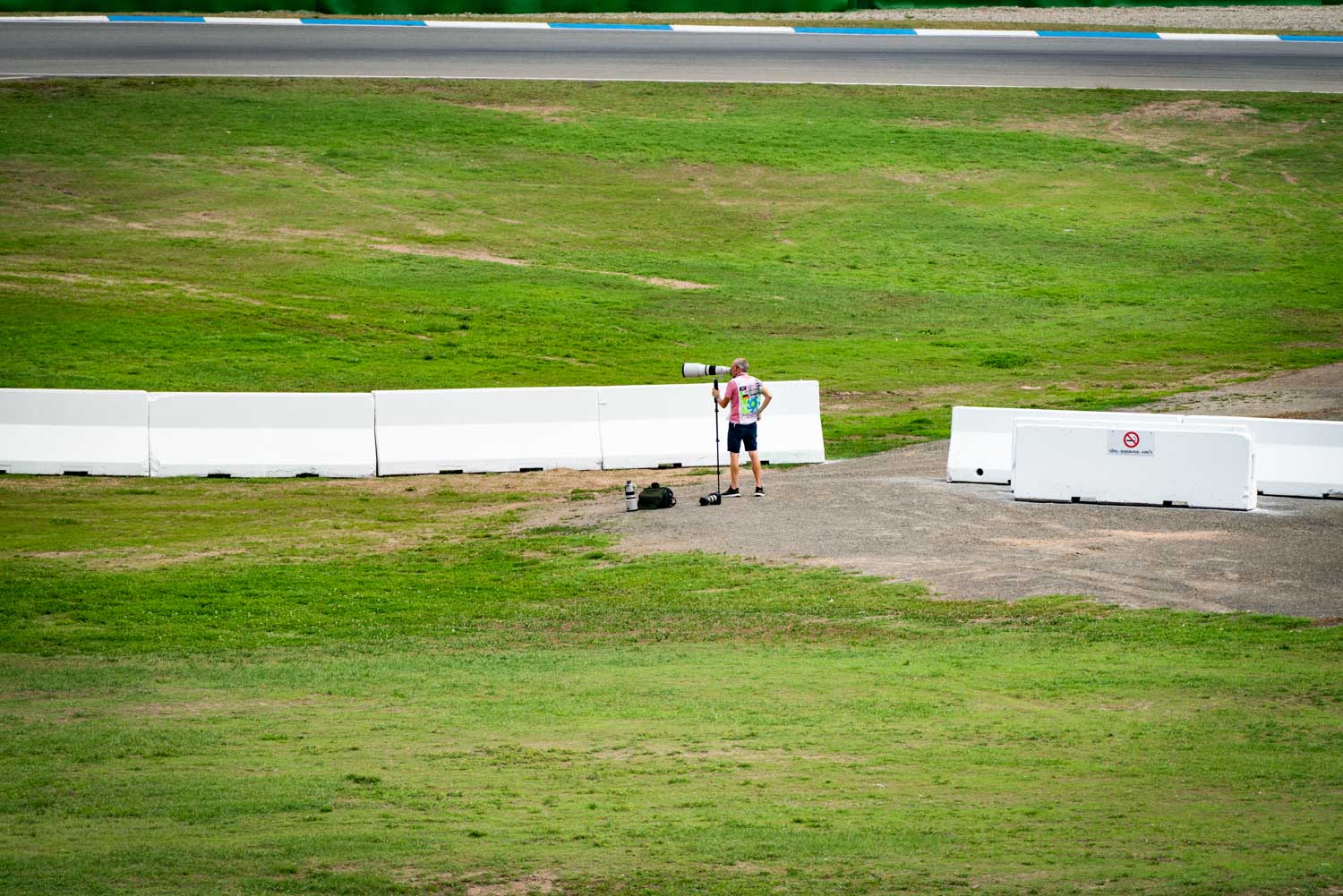 Photographer with a large camera lens stands near a grassy trackside, capturing a motor racing event.