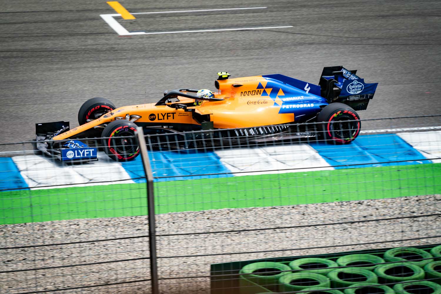 Orange and blue Formula 1 race car on track, sponsored by Lyft, with blurred motion and surrounding safety barriers.