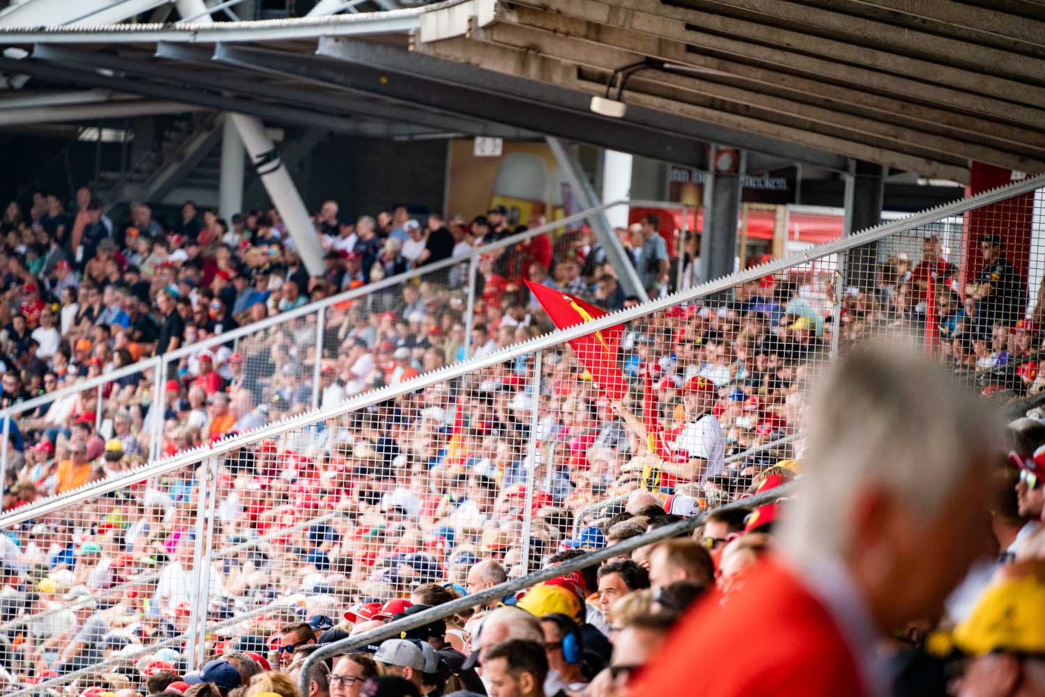 Crowded sports stadium with fans wearing red and waving flags; metal fencing in foreground.