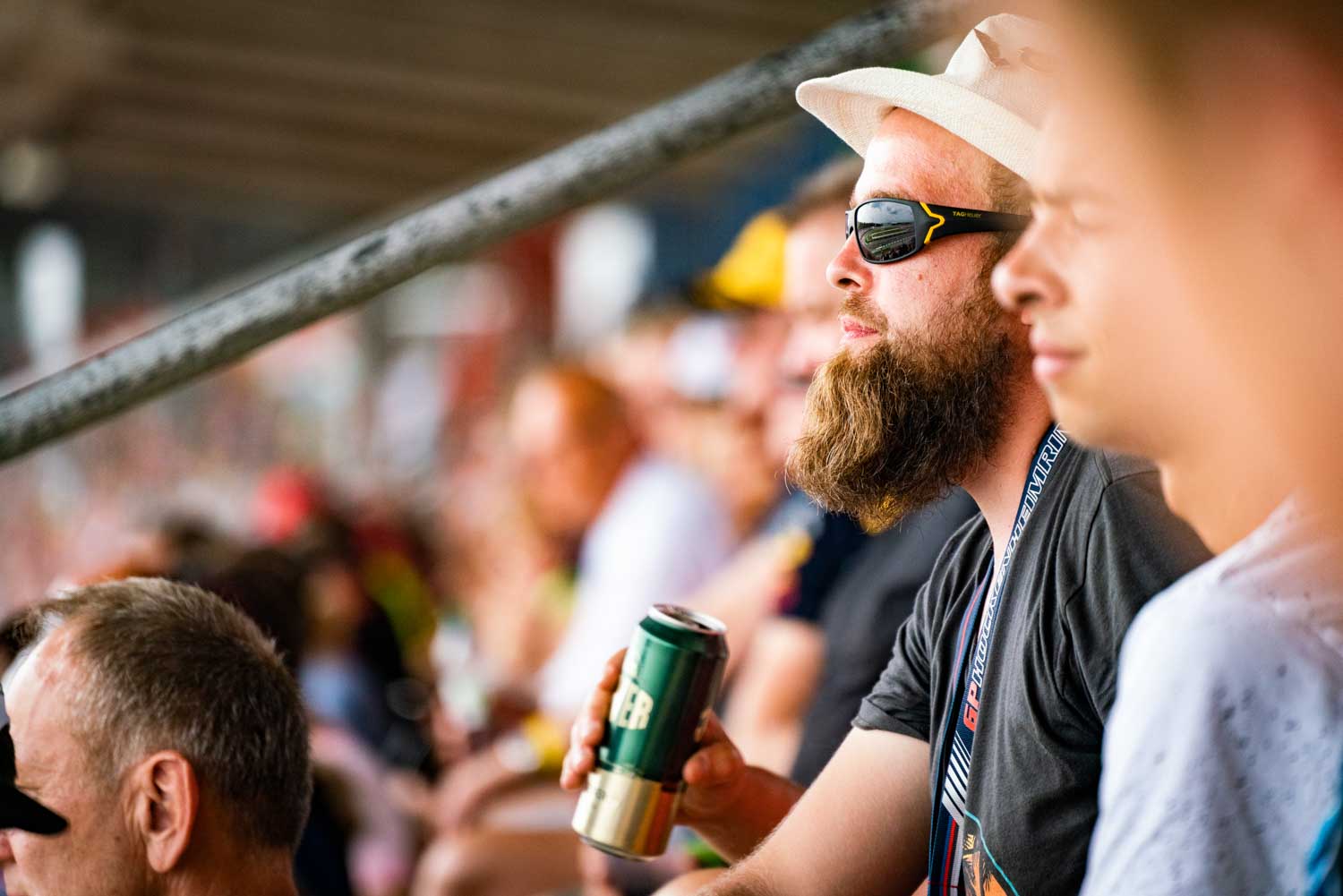 Bearded man in sunglasses and hat holds drink, watching event among a diverse crowd in stadium seating.
