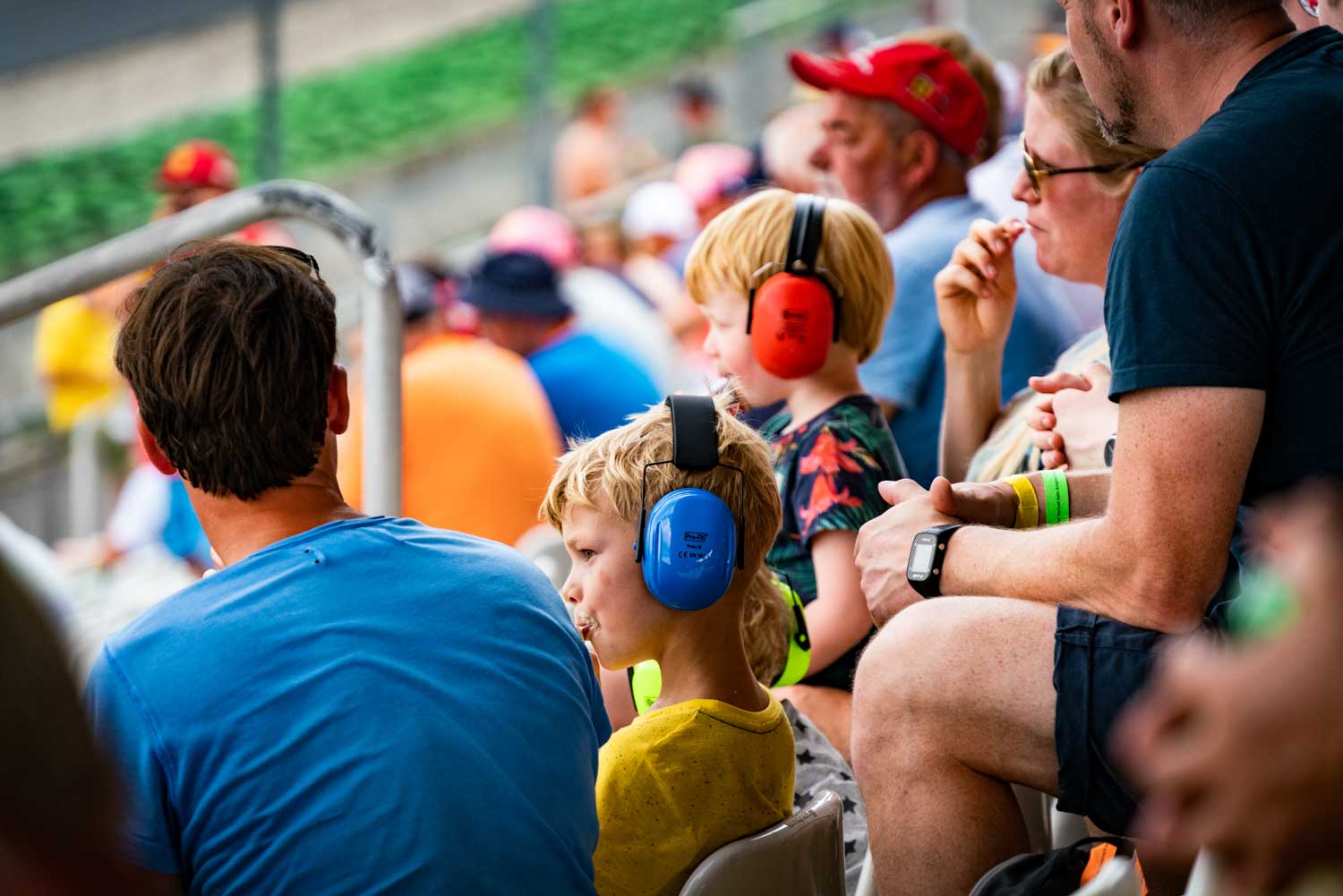 Spectators at a racetrack, including children with protective earmuffs, watching an event from the stands.