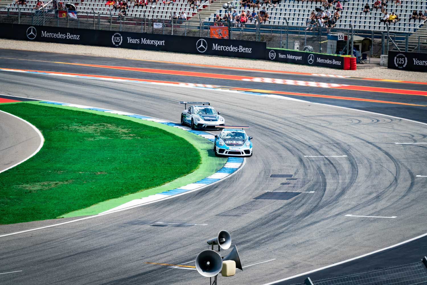 Two race cars on a track taking a sharp turn, with spectators in the grandstands and Mercedes-Benz signage visible.