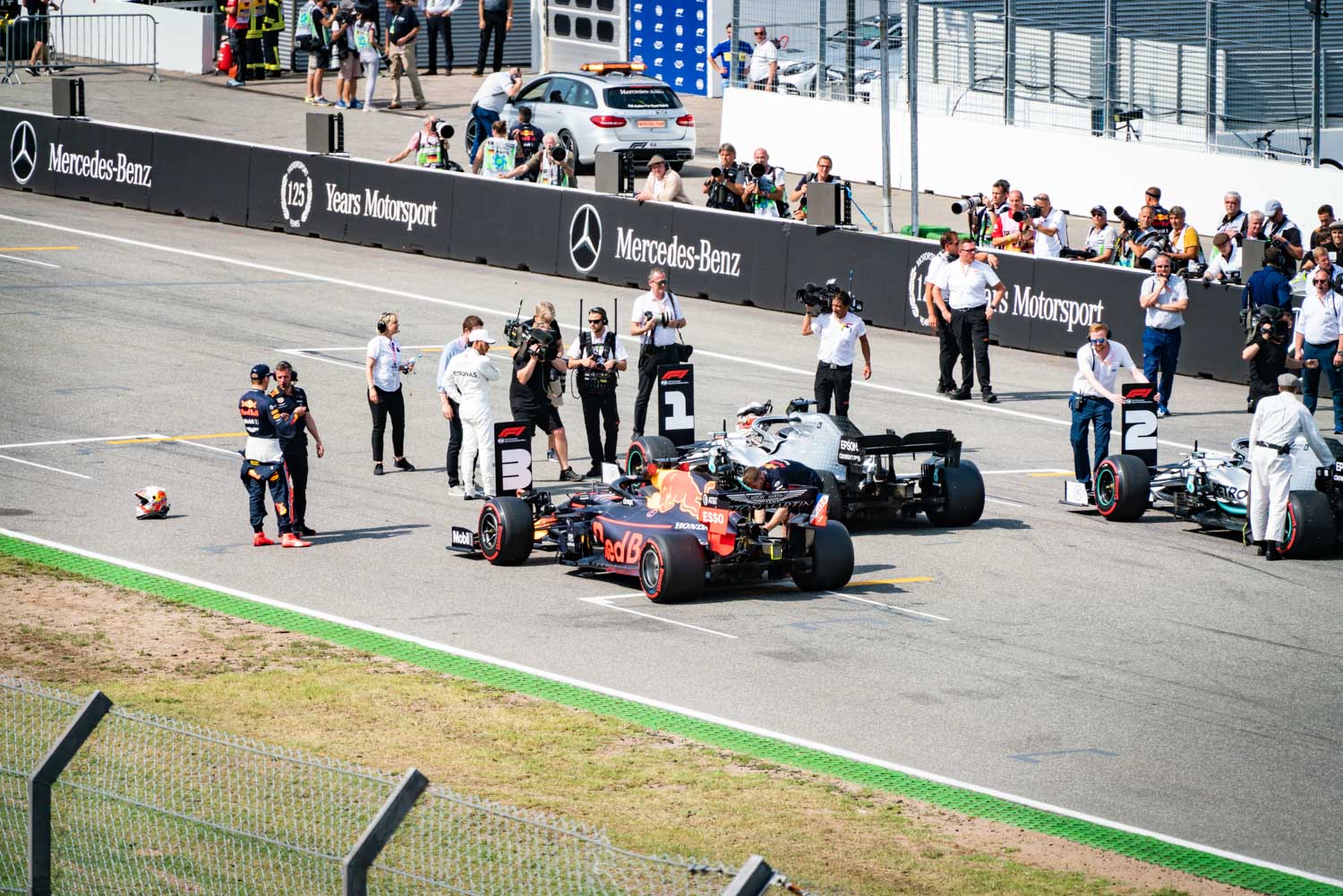 Formula 1 drivers and cars on starting grid, surrounded by team members and media at a racing event.