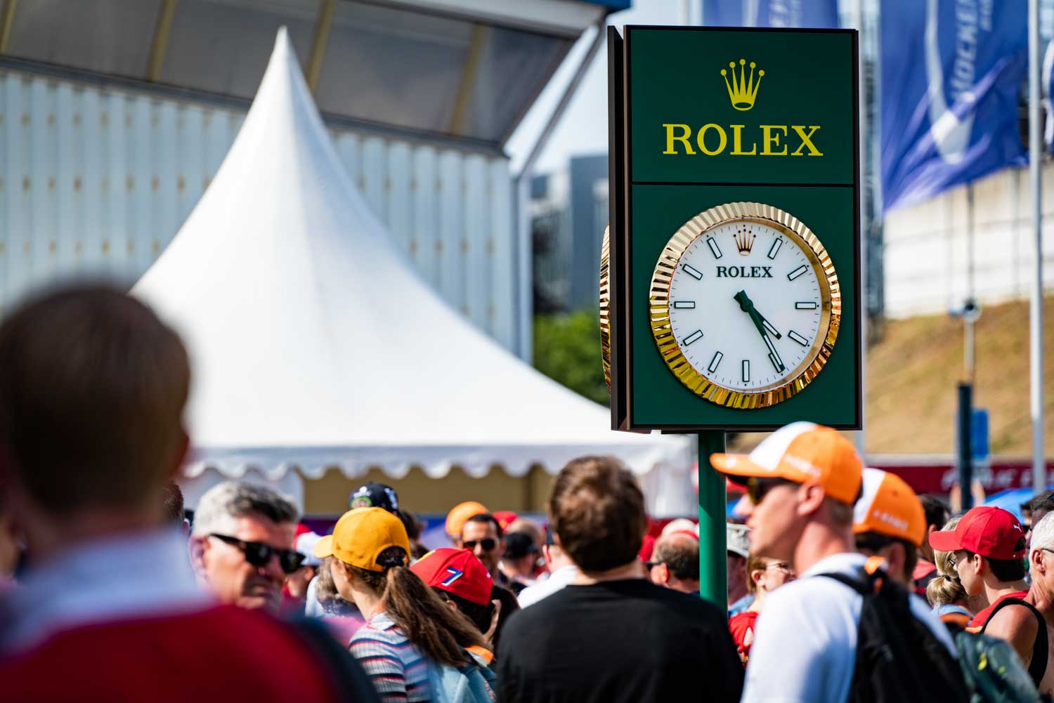 Crowd at an outdoor event near a large Rolex clock display under a white tent.