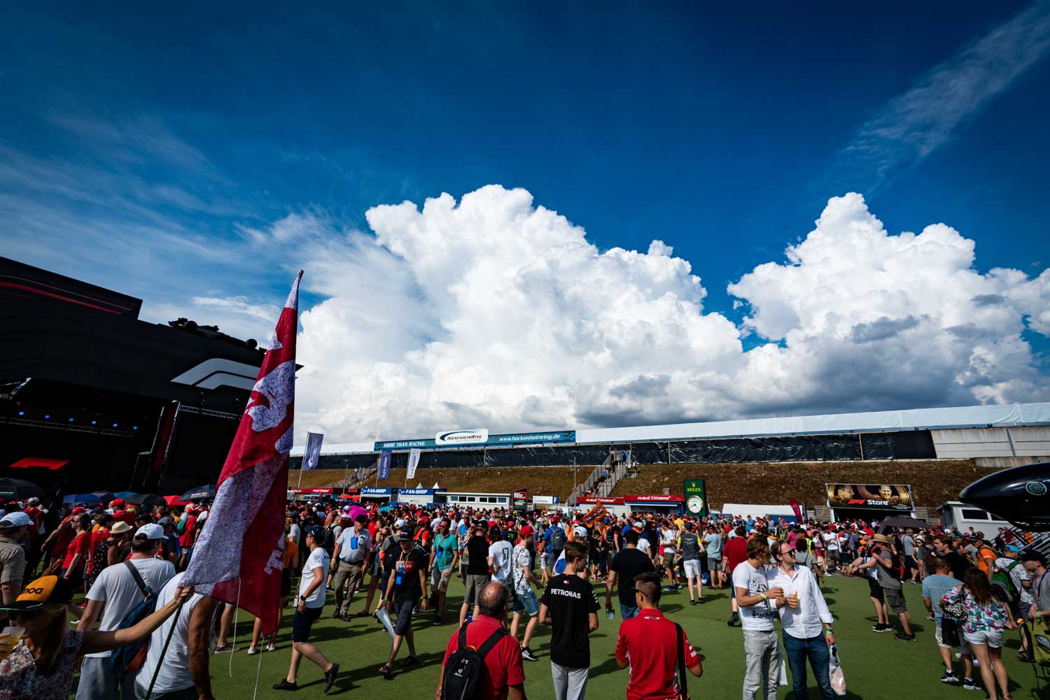 Crowd at a motorsport event under a sunny sky with fluffy clouds, featuring flags and a stage in the background.