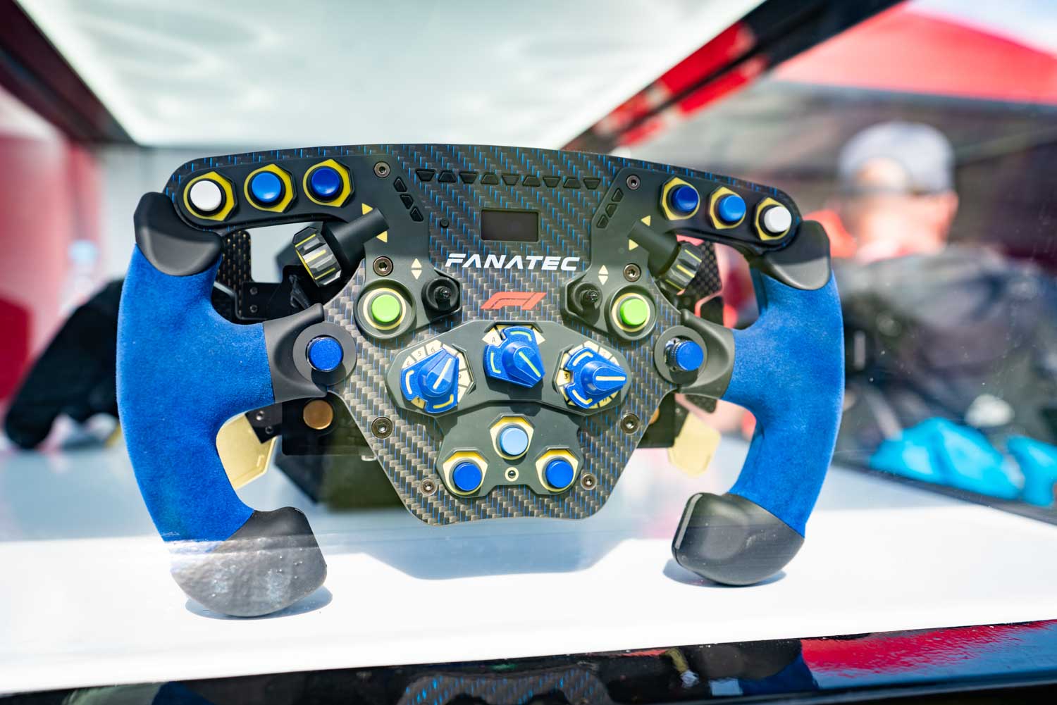 Close-up of a Fanatec racing wheel with blue handles and multiple buttons, showcased on a white surface in bright lighting.