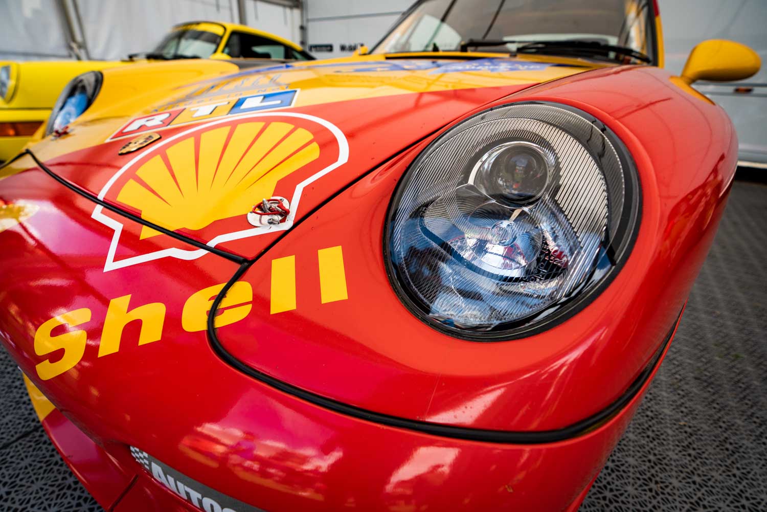 Close-up of a red Porsche race car with Shell and RTL logos on the hood, parked indoors next to a yellow car.