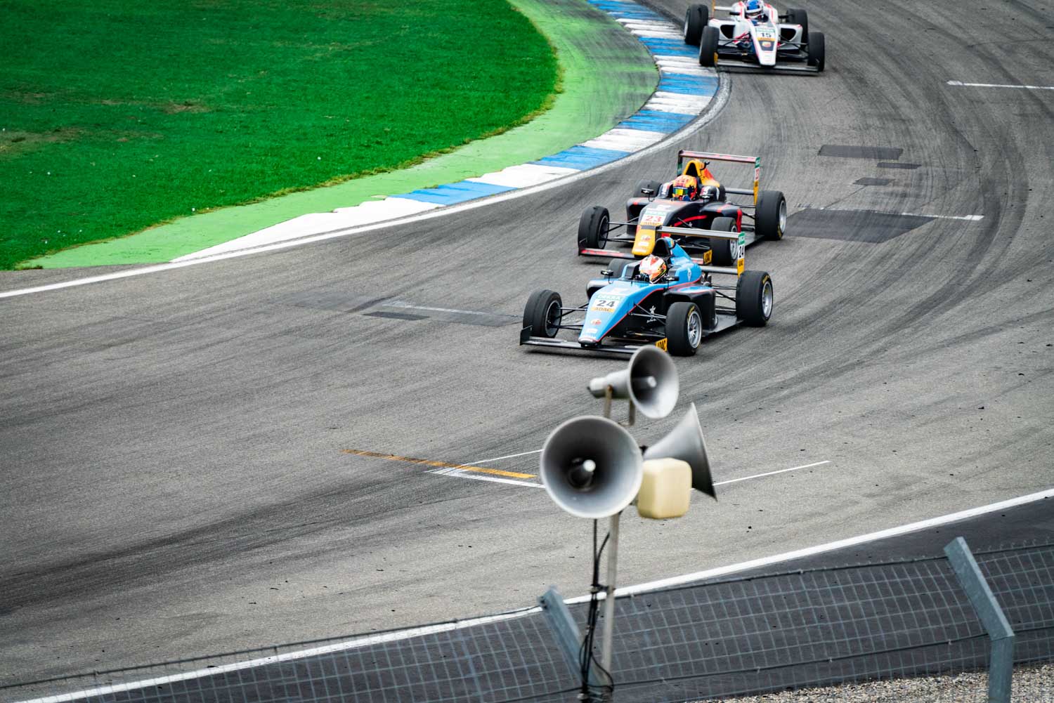 Race cars on track rounding a bend, with grass and grandstand background, during a motorsport competition.