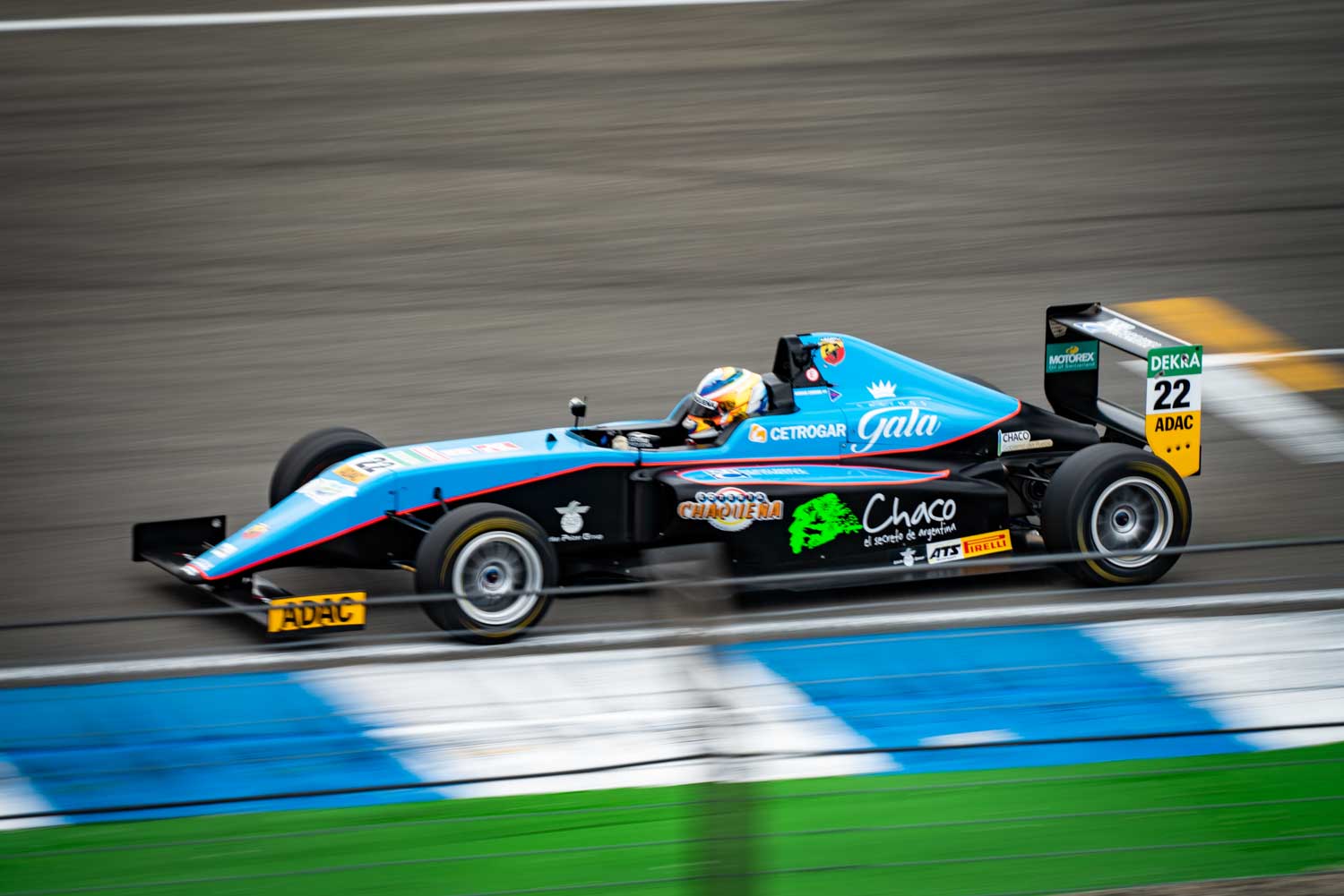 Blue Formula 4 race car speeding on the track, showcasing sponsorship logos and driver in helmet.