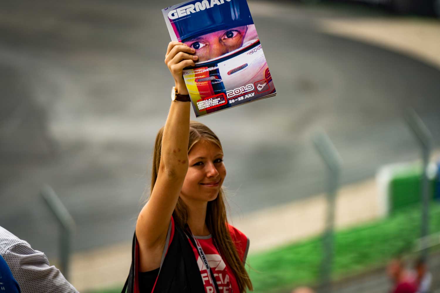Smiling woman at a racetrack holding a German Grand Prix program booklet with a driver’s face on the cover.