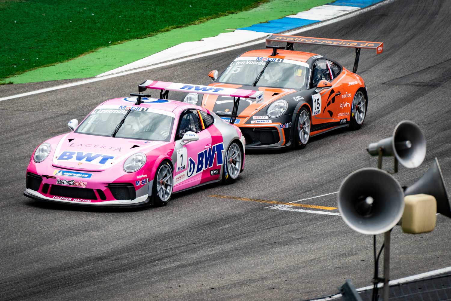 Two Porsche racing cars, one pink and one orange, speed around a racetrack corner during a competitive motorsport event.