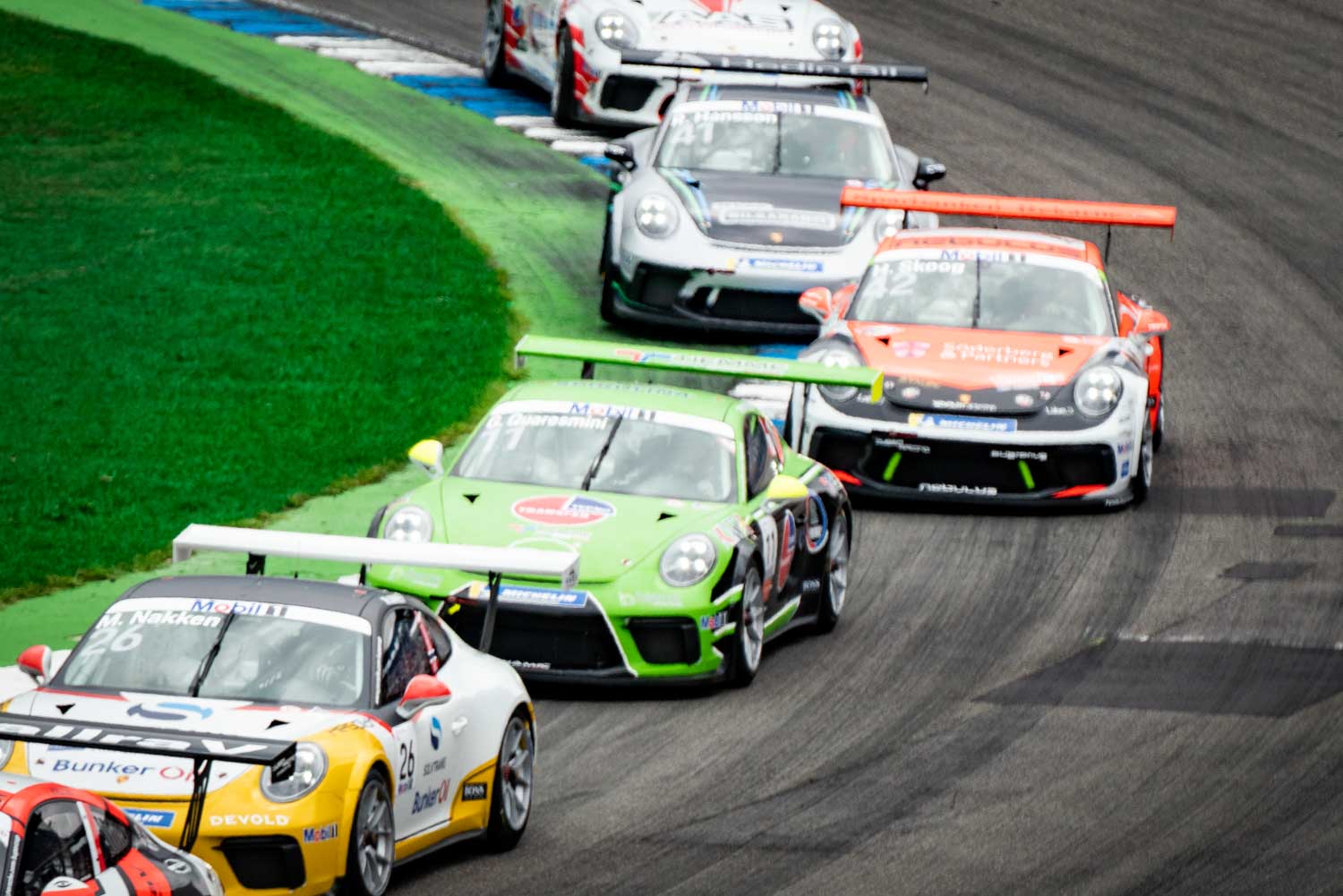 Race cars speeding on track during competition, green car leading. Racing action on a curving circuit with cheering spectators.