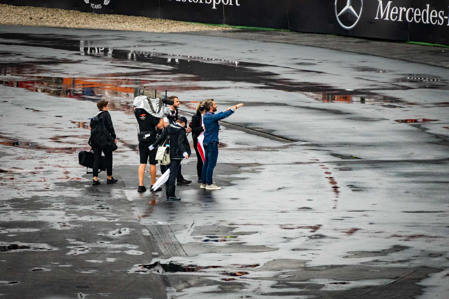 People stand on a wet racetrack, engaged in discussion, with logos in the background.