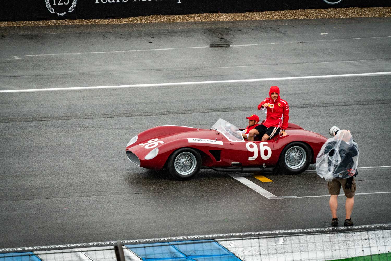 Vintage red race car number 96 on wet track, two people inside in red clothing, photographed by a person with a camera.