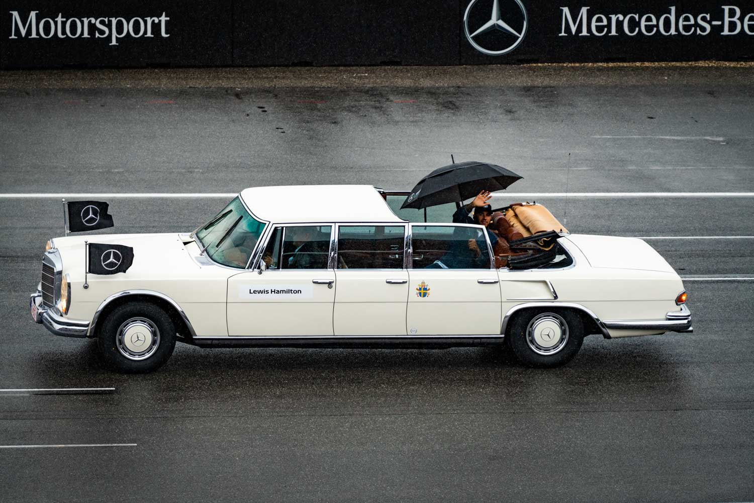 White Mercedes limousine with flags on a wet track, passenger holding umbrella.