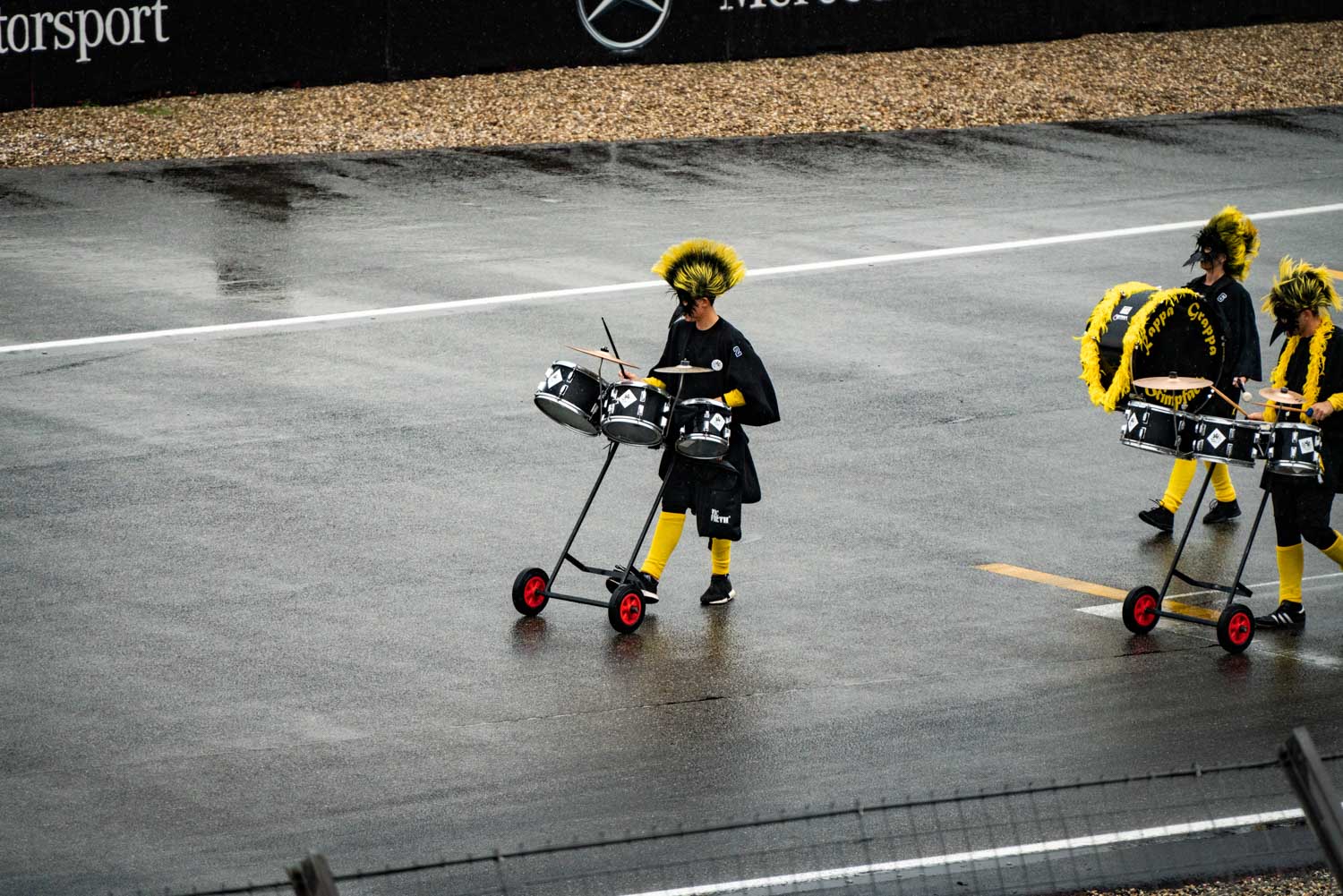 Drummers in yellow-black costumes perform on a wet track at an outdoor event.