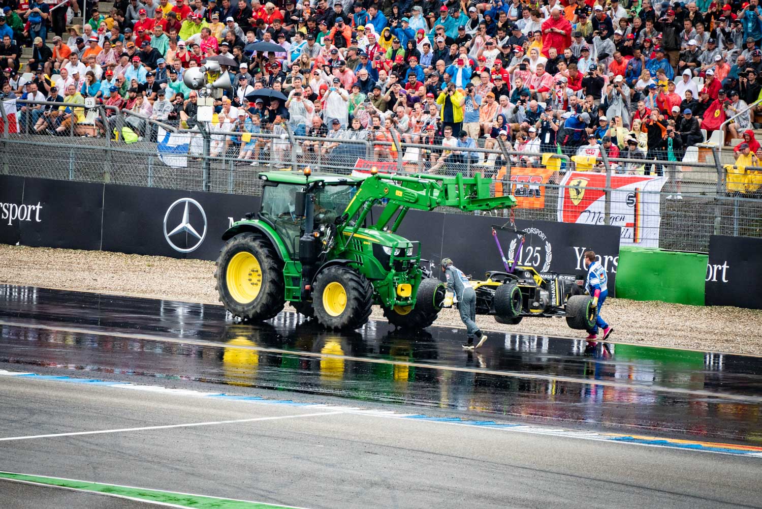 Green tractor removing a race car from a wet track during a motorsport event, with a crowd in the background.